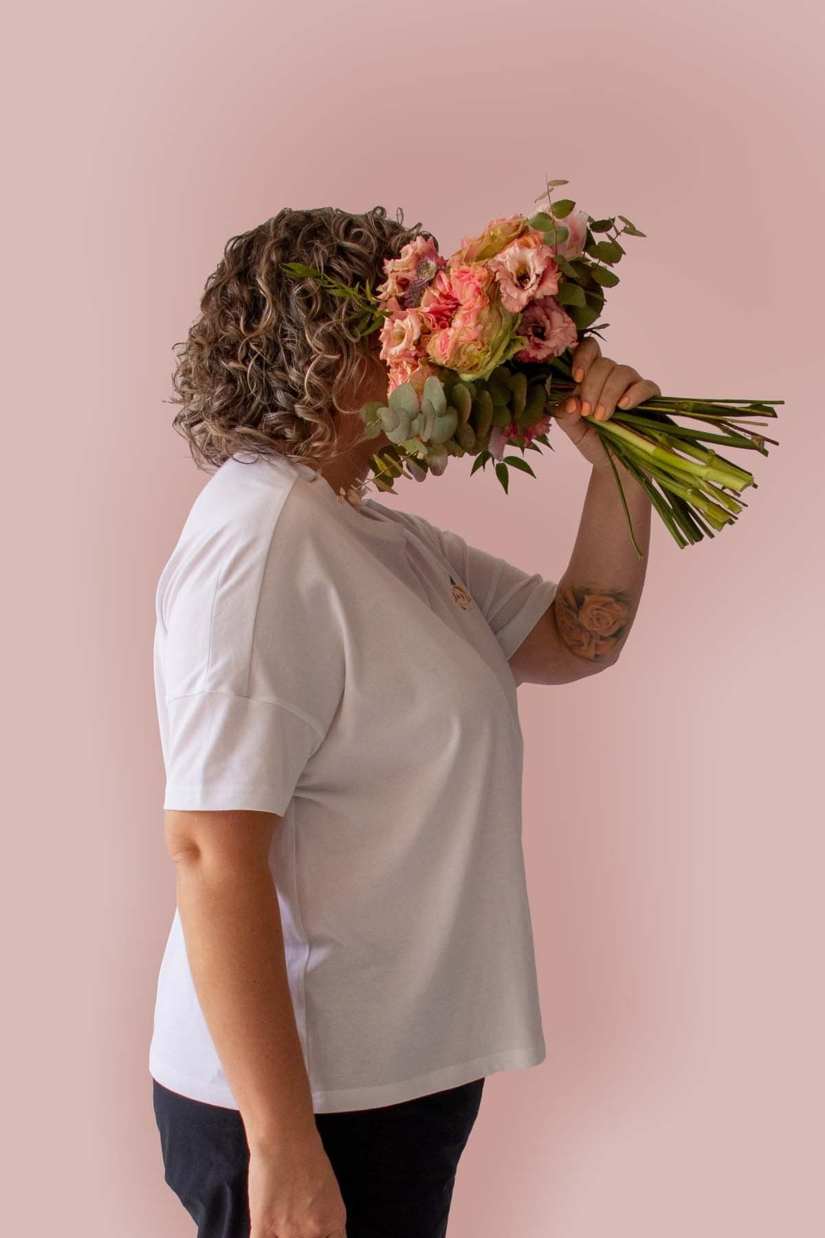 Side view of Asmuss Boxy T-shirt in White Organic Cotton and Recycled Polyester with Embroidered Rose detail worn by Clare holding a beautiful bunch of flowers, that echo the embroidery, in front of her face.