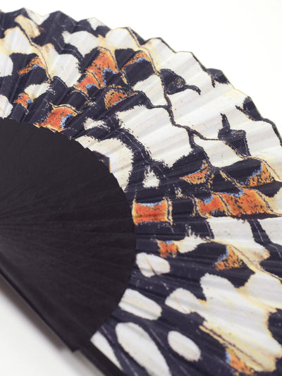 Close up of Khu Khu Black Nymph Hand Fan with black sticks and a symmetrical butterfly print in black, off white and hints of blue and burnt orange.