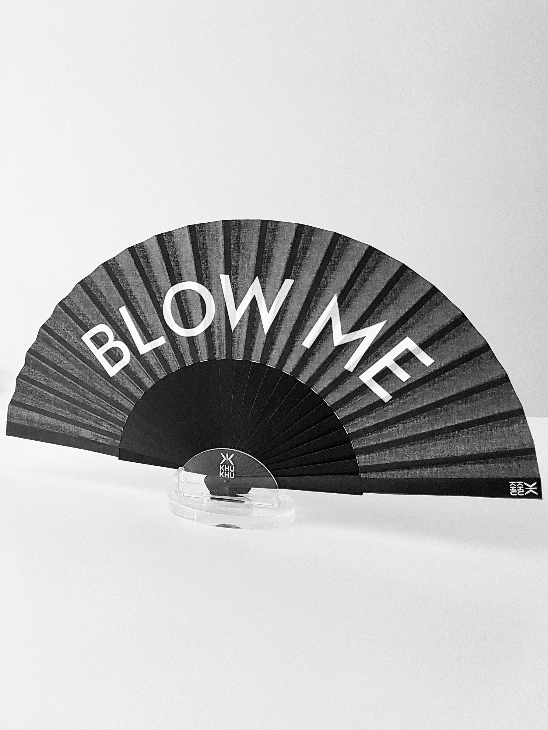 Khu Khu Blow Me Hand-Fan. White Font BLOW ME on black background with black wooden sticks and black rivet. Fan sits in acrylic table stand. 